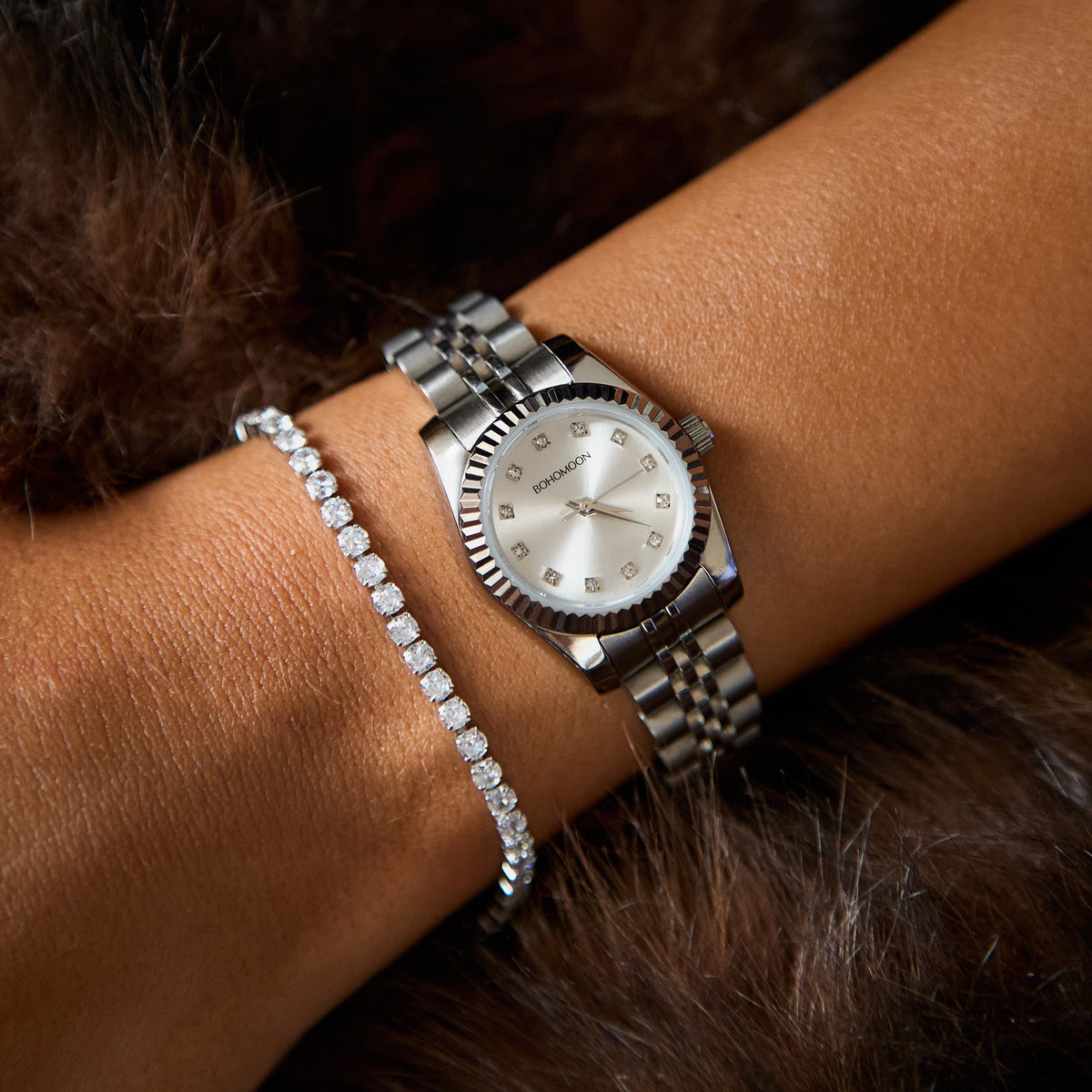A close-up of a woman's wrist wearing a silver-tone Swatch watch with a mother-of-pearl dial and a matching silver bracelet, alongside a diamond-set bracelet, set against dark brown fur.