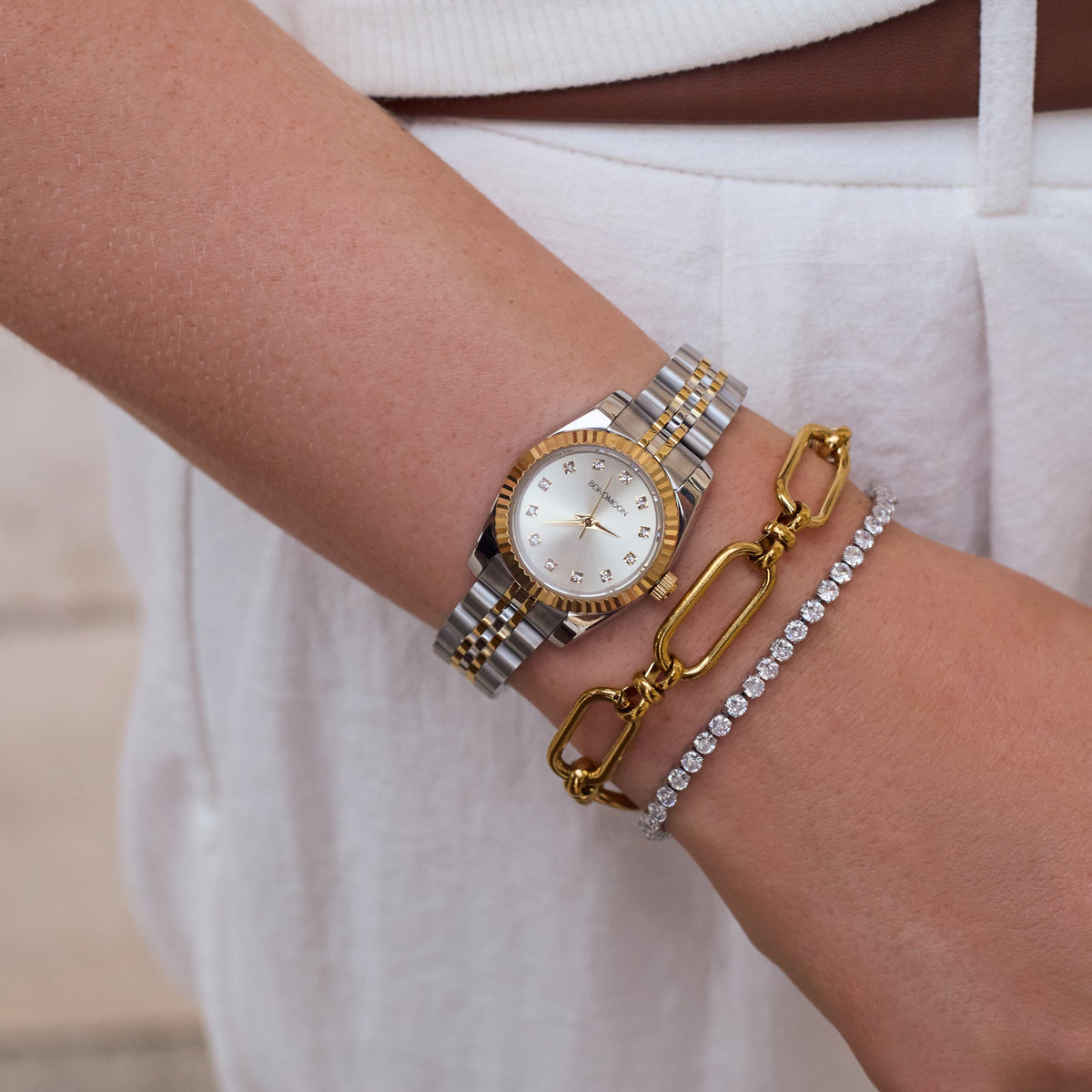 A close-up of a person's wrist wearing a silver and gold two-tone watch, layered with a gold chain link bracelet and a silver tennis bracelet.